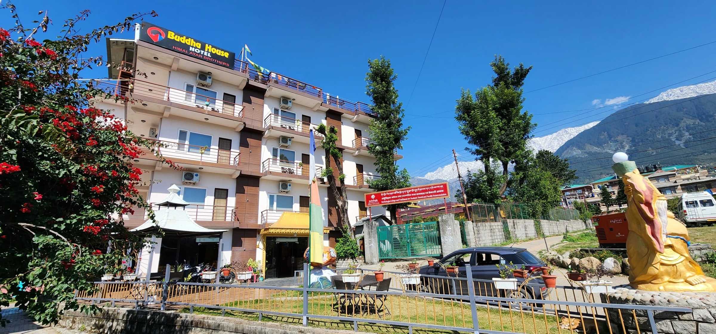 Hotel Buddha House: a five-storey building with balconies and its rooftop sign, a garden in front and snow on the Dhauladhar behind