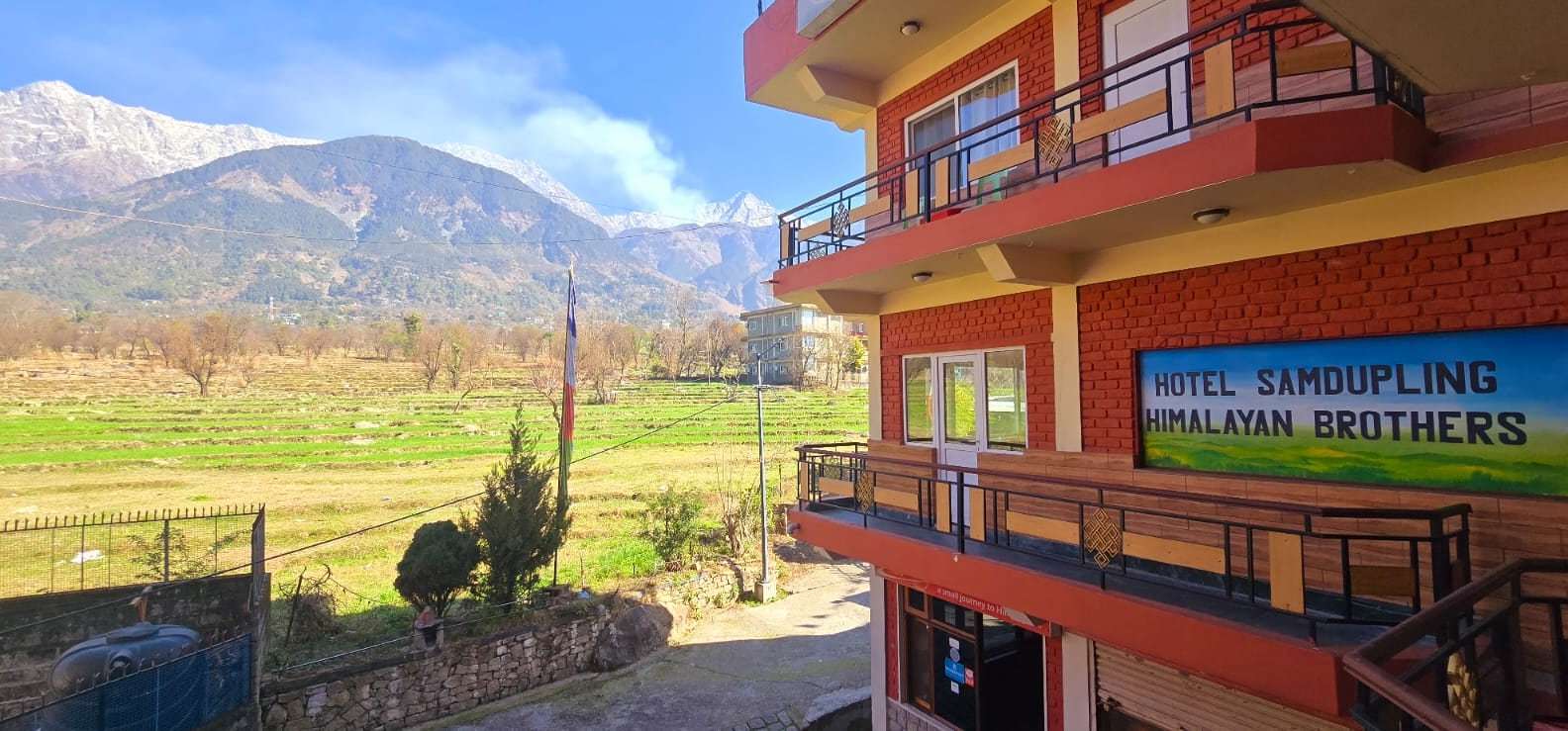 Hotel Samdupling’s red-brick front and balconies, with green fields and the Dhauladhar behind