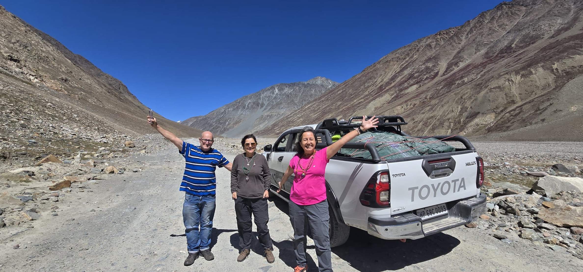 Three of our guests wave beside a pickup on a gravel road between bare mountains in Spiti