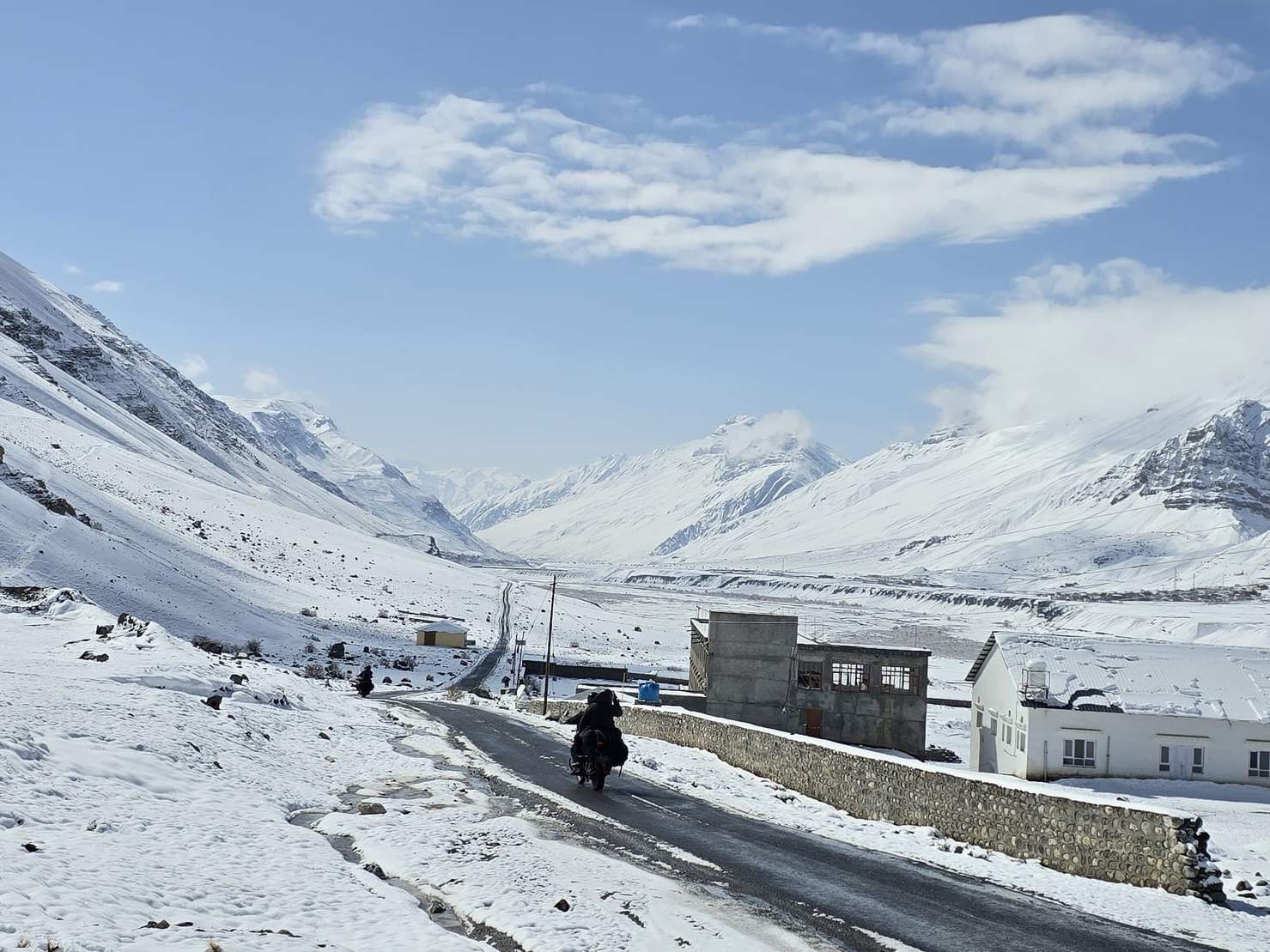 A motorcyclist rides a cleared road through snow near Kaza, with white mountains under a blue sky
